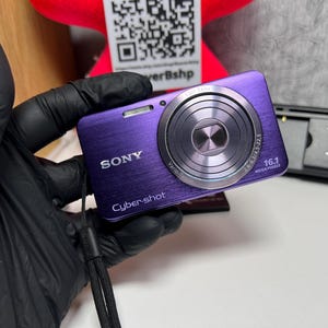 May include: A purple Sony Cyber-shot digital camera with a brushed metal finish, featuring a Carl Zeiss Vario-Tessar lens, a 16.1 megapixel sensor, and an attached black wrist strap. The camera body displays the Sony and Cyber-shot logos.
