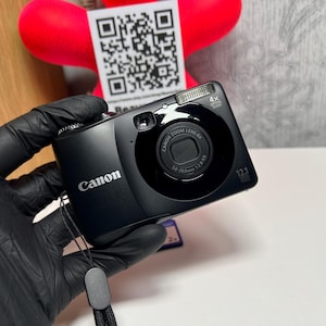 Könnte beinhalten: Eine schwarze Canon Digitalkamera mit 4-fach optischem Zoomobjektiv, 12,1-Megapixel-Sensor und integriertem Blitz. Auf der Vorderseite befinden sich das Canon-Logo, die Objektivspezifikationen 5,0-20,0mm 1:2,8-5,9 und eine schwarze Handschlaufe.