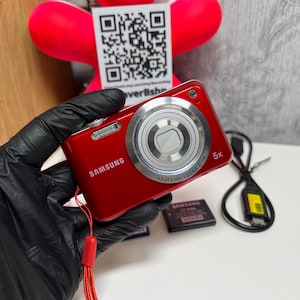 Op de afbeelding: Een rode Samsung digitale camera met een 5x optische zoomlens, een zilverkleurige lenscilinder en een rood polsbandje. De camerabehuizing toont het Samsung-logo aan de voorzijde, vergezeld van een zwarte accu en een zwarte USB-oplaadkabel.