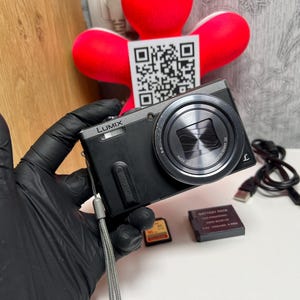 Op de afbeelding: Een zwarte Panasonic Lumix digitale camera met een zilveren lensring en een grijze polsband. Inbegrepen zijn een zwarte accu, een kleine geheugenkaart en een zwarte USB-oplaadkabel. De camera is voorzien van een opvallende lens.