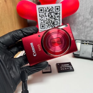 Może przedstawiać: Metaliczny czerwony aparat cyfrowy Canon IXUS z obiektywem z 10-krotnym zoomem, wbudowaną lampą błyskową i czarnym paskiem na nadgarstek. W zestawie znajduje się czarna ładowarka, czarny akumulator oraz mała karta pamięci.