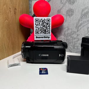 Puede incluir: Videocámara Canon LEGRIA HF S21 negra con un lector de tarjetas blanco, una tarjeta SD azul de 8 GB y dos cargadores de batería negros. Una figura roja con un código QR en el fondo.