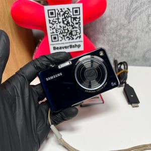 May include: A dark blue Samsung digital camera featuring a 5x optical zoom lens with 4.9-24mm 1:3.5-5.9 27mm specifications printed on the front. The compact device includes a built-in flash, a wrist strap, and a black USB charging cable.