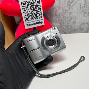 May include: A silver Olympus FE-210 digital camera featuring a compact body, a circular lens housing, a built-in flash, and a grey wrist strap. The front of the camera displays the Olympus brand name and the model number FE-210 clearly.