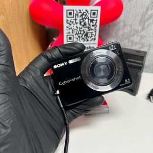 May include: A black Sony Cyber-shot digital camera featuring an 8.1 megapixel Carl Zeiss Vario-Tessar lens, a built-in flash, and a black wrist strap. The camera body displays the Sony and Cyber-shot logos on the front face.