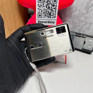 May include: A silver Samsung i85 digital camera with a brushed metal finish, featuring a 5X optical zoom lens, a built-in flash, and a wrist strap. The camera body displays the Samsung brand name, lens specifications, and the model number i85.
