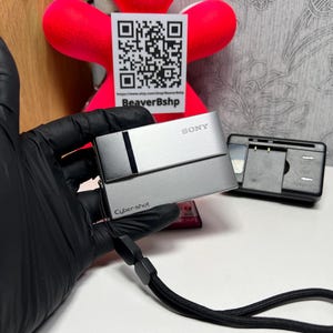 May include: A silver Sony Cyber-shot digital camera with a black wrist strap is held in a black gloved hand. A black camera charger sits to the right. The background includes a red plush toy and a QR code.