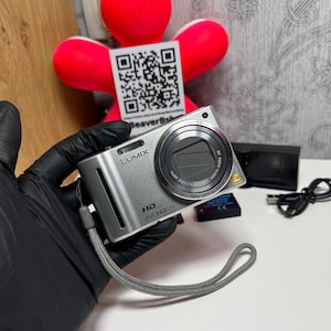 Op de afbeelding: Een zilverkleurige Panasonic Lumix digitale camera met een Leica DC Vario-Elmar lens met 12x optische zoom, HD AVCHD Lite-branding en een grijze polsband. De camerabehuizing bevat een ingebouwde flitser, een ontspanknop en een klein gouden L-logo.