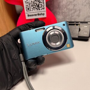 Op de afbeelding: Een metallic blauwe Panasonic Lumix digitale camera met een Leica DC Vario-Elmarit lens met Mega O.I.S. technologie, een zilveren polsbandje en een klein goudkleurig L-logo op de voorzijde, vergezeld door een zwarte acculader.