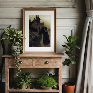 May include: A framed artwork depicting a figure walking on a path near a house, in a muted color palette. The artwork is displayed on a wooden console table with drawers, alongside potted plants and a terracotta pot. The scene is set against a neutral wall.
