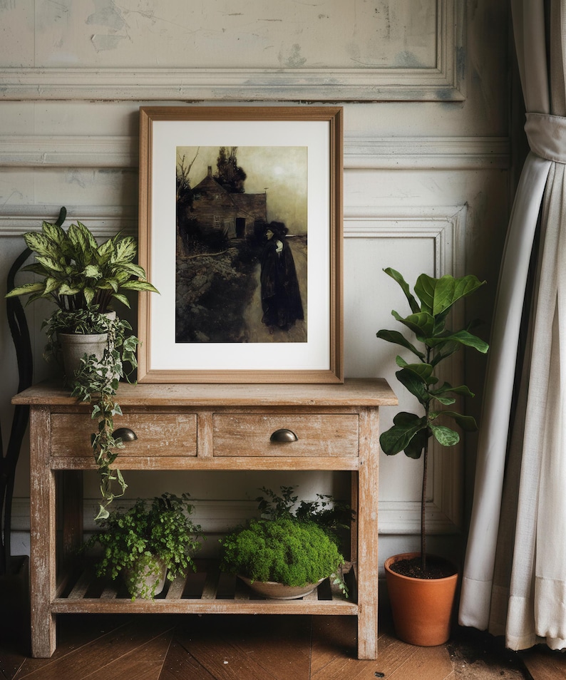 May include: A framed artwork depicting a figure walking on a path near a house, in a muted color palette. The artwork is displayed on a wooden console table with drawers, alongside potted plants and a terracotta pot. The scene is set against a neutral wall.