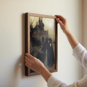 May include: A framed print of a dark, moody landscape featuring a house and a figure on a path. The artwork is in a wooden frame, and the overall aesthetic is vintage. The print is being held up against a white wall.