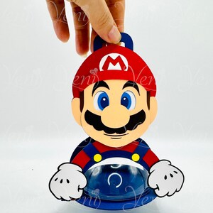 Puede incluir: Decoraci&oacute;n de personaje de Mario de estilo caricatura. La figura tiene un sombrero rojo con una "M" blanca, ojos azules, un gran bigote negro y un atuendo azul y rojo. La decoraci&oacute;n est&aacute; sujeta por una mano.