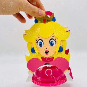 Puede incluir: Recorte de papel de la Princesa Peach de la serie de videojuegos Super Mario. El personaje tiene cabello amarillo, ojos azules y un vestido rosa. Lleva una corona dorada con una joya roja y sostiene un objeto rosa transparente.