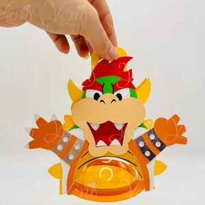 Puede incluir: Decoraci&oacute;n de papel artesanal con tem&aacute;tica de Bowser, colorida. El dise&ntilde;o presenta un Bowser de dibujos animados con detalles en naranja, amarillo, rojo y verde. La decoraci&oacute;n tiene una abertura circular y est&aacute; sujeta por una mano.