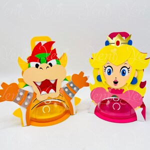 Puede incluir: Dos recortes de papel hechos a mano y coloridos de personajes de videojuegos. Uno es Bowser, con detalles naranjas, amarillos, rojos y verdes. La otra es la Princesa Peach, con cabello amarillo, un vestido rosa y una corona. Ambos tienen recipientes de pl&aacute;stico transparente.