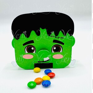 May include: A green, Frankenstein-themed paper box with black hair and stitched details. The box has a smiling face with pink cheeks and brown eyes. Colorful candies are scattered in front of the box. The image also includes download icons.