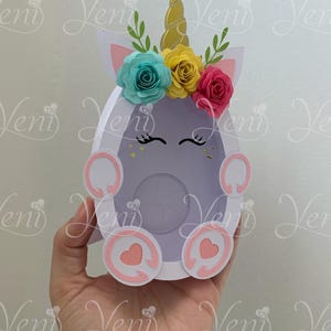 Puede incluir: Una manualidad de papel en forma de unicornio blanco con un cuerno dorado, orejas rosas y una corona de flores con flores azules, amarillas y rosas. El unicornio tiene corazones rosas en sus patas y un fondo blanco.