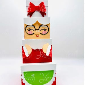 May include: A stack of four decorative boxes, each with a different design. The top box has a red bow, the second box features a face with glasses, the third box has white and beige details, and the bottom box is green and white.