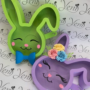 Puede incluir: Dos alcancías con forma de conejo, una verde con un lazo azul y la otra morada con un adorno floral. Ambas tienen una ventana de plástico transparente para monedas.