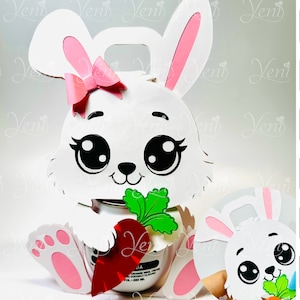 Puede incluir: Caja de regalo en forma de conejo blanco con un lazo rosa, ojos grandes y una zanahoria. La caja está diseñada para contener un pequeño regalo o golosina. La imagen incluye texto en español e inglés, indicando que el archivo es para Silhouette.