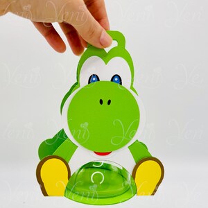 Puede incluir: Un recorte de papel verde con tem&aacute;tica de Yoshi, con detalles blancos, ojos azules y boca roja. El recorte tiene un lazo en la parte superior y un elemento transparente en forma de c&uacute;pula en la parte inferior. El dise&ntilde;o recuerda a un personaje popular de videojuegos.