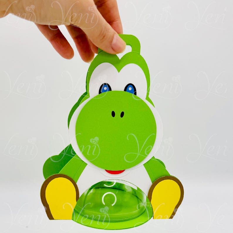 Puede incluir: Un recorte de papel verde con tem&aacute;tica de Yoshi, con detalles blancos, ojos azules y boca roja. El recorte tiene un lazo en la parte superior y un elemento transparente en forma de c&uacute;pula en la parte inferior. El dise&ntilde;o recuerda a un personaje popular de videojuegos.