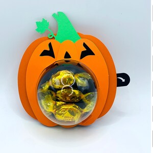 Puede incluir: Calabaza de papel naranja y verde con una cúpula de plástico transparente llena de caramelos de chocolate. La calabaza tiene una cara de calabaza negra. La imagen incluye texto para "ZIP", "VIDEO", "STUDIO", "SVG" y "Iris Shutter".