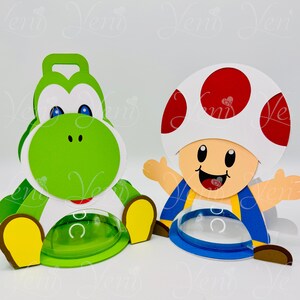 Puede incluir: Dos recortes de personajes coloridos de estilo caricatura. Uno es una figura verde de Yoshi con vientre blanco y detalles amarillos. El otro es un personaje de Toad con un gorro de hongo blanco con lunares rojos, chaleco azul y detalles amarillos. Ambos tienen recipientes de pl&aacute;stico transparente.