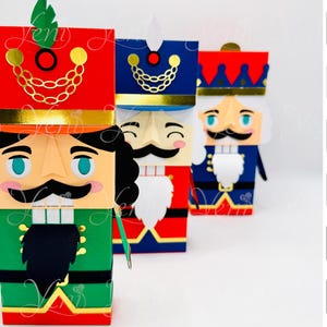 Puede incluir: Tres figuras de cascanueces de papel de colores. El cascanueces delantero es verde, rojo y negro, con una cadena dorada y un bigote negro. Los otros dos son rojos y azules, con detalles dorados. La imagen incluye texto: "Jem", "ZIP", "VIDEO", "STUDIO", "SVG", "Money Box".