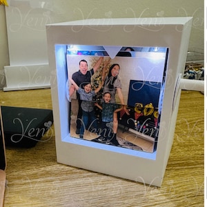 May include: A white, illuminated photo display box with a family photo inside. The box has a rectangular shape with a cutout to showcase the photo. The photo features a family of four. The box has the word "Veni" in a script font.