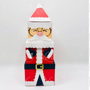 Op de afbeelding: Een stapel van drie decoratieve dozen die op de kerstman lijken. De bovenste doos is een rode en witte hoed, de middelste doos is het gezicht van de kerstman met een bril en de onderste doos is zijn pak met een gouden gesp. Voor Silhouette.