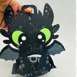 Könnte beinhalten: Ein schwarzer Papierdrache mit großen grünen Augen und weißen Akzenten. Der Drache hat einen Griff oben und Flügel an den Seiten. Das Gesicht des Drachen ist der Mittelpunkt, mit ausgeschnittenen Augen und Details. Der Drache wird von einer Hand gehalten.