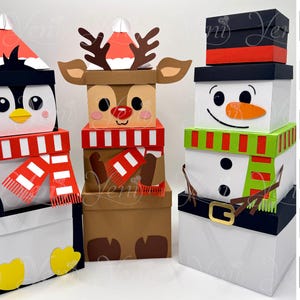 Può includere: Tre scatole impilate a tema natalizio: un pinguino, una renna e un pupazzo di neve. La scatola del pinguino è bianca e nera con una sciarpa rossa e bianca. La scatola della renna è marrone con corna e una sciarpa rossa. La scatola del pupazzo di neve è bianca con una sciarpa verde e rossa.