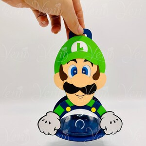 Puede incluir: Una manualidad de papel del personaje Luigi de la serie Super Mario Bros. El personaje tiene un gorro verde con una "L" blanca, un gran bigote negro y ojos azules. El personaje sostiene un recipiente azul.