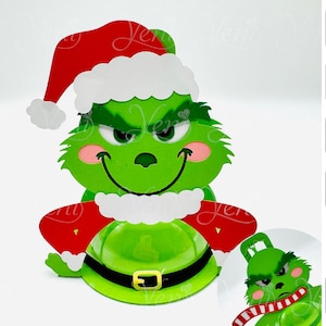 Puede incluir: Un personaje de dibujos animados verde con un gorro de Papá Noel rojo y un borde blanco. El personaje tiene una sonrisa traviesa y lleva un atuendo verde con un cinturón negro. Diseño digital.