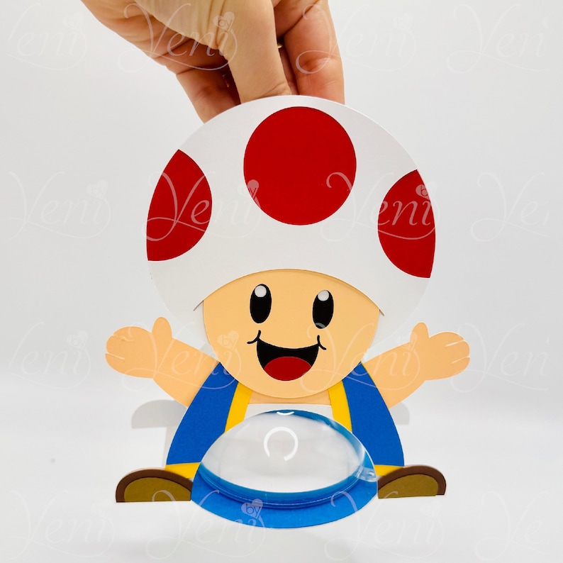 Puede incluir: Una figura de papel de un personaje de dibujos animados con una gran gorra de hongo blanca con lunares rojos. El personaje tiene una cara sonriente, un mono azul y los brazos extendidos. Debajo hay un objeto transparente en forma de c&uacute;pula.