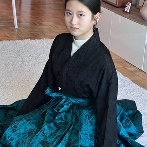 以下が含まれることがあります： 伝統的な韓国のハンボクを着た人。この衣装は、花の刺繍が施された黒いジャケットと、花柄のターコイズ色のスカートが特徴です。スカートはターコイズ色のリボンで結ばれています。