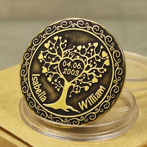 Peut inclure: Une pièce de monnaie en métal couleur or avec un motif d'arbre comportant un cœur central avec la date 04.06.2003. Les noms "Isabella" et "William" sont inscrits de chaque côté de l'arbre.