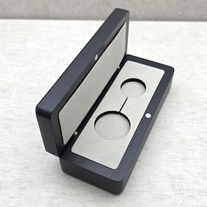 May include: A rectangular black wooden display box with a hinged lid, featuring a light gray interior lining with two circular cutouts designed to hold small round items. The box has a smooth finish and small magnetic closures on the front edge.
