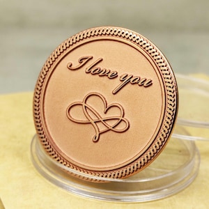 May include: A rose gold coin with a decorative border and the words "I love you" engraved on the front. The coin also features a stylized infinity symbol with a heart in the center.