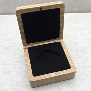May include: A square wooden box with a light brown wood grain finish and a hinged lid. The interior is lined with black velvet, featuring a central circular cutout in the base for holding an item. The box includes a small magnetic closure.