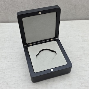 May include: A square, dark wood display box with a hinged lid and a magnetic closure. The interior features a light grey, soft-touch lining with a central, cloud-shaped cutout designed to hold a small item securely in place.