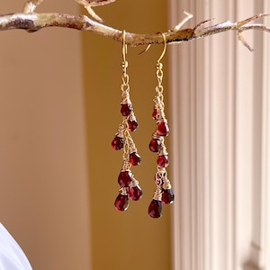 May include: Dangling earrings with a gold-colored wire and chain design. Each earring features multiple teardrop-shaped, deep red gemstones. The earrings are displayed against a soft, neutral background, highlighting their elegant design.