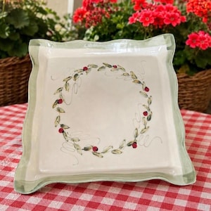 May include: A square ceramic plate with a light green painted border and a central hand-painted wreath design. The wreath consists of thin green leaves, delicate swirling vines, and several small red ladybugs scattered along the circular pattern.