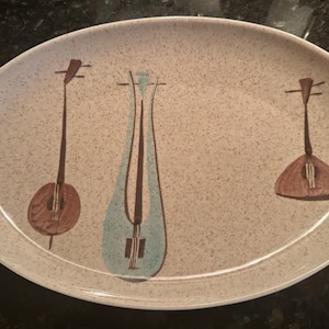 May include: Oval serving platter with a speckled beige surface. The platter features three stylized musical instrument designs in brown and teal. A decorative piece for serving food.