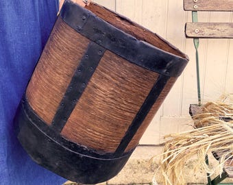 May include: A cylindrical wooden bucket featuring a dark brown wood grain texture, reinforced with wide black metal bands at the top, bottom, and vertically along the sides, secured with visible metal rivets throughout the structure.