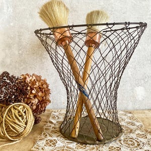 Può includere: Un cestino di filo metallico vintage contiene due pennelli con manici in legno e ghiera in rame. I pennelli hanno setole naturali. Una palla di spago e fiori secchi sono accanto al cestino. Il cestino è su una tovaglietta di pizzo.