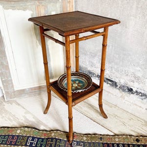 May include: A vintage bamboo side table with a square top and a lower shelf. The table has a natural wood finish and a woven top and shelf. A decorative plate with a floral design rests on the shelf. The table is approximately 71 cm tall.