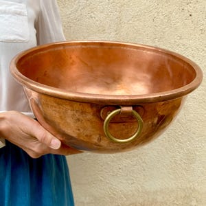 Pode incluir: Uma taça grande e redonda de cobre polido com uma superfície lisa e refletora. A taça apresenta um rebordo espesso e arredondado e uma pega em forma de anel de latão fixada na lateral com uma placa metálica retangular de tom quente.